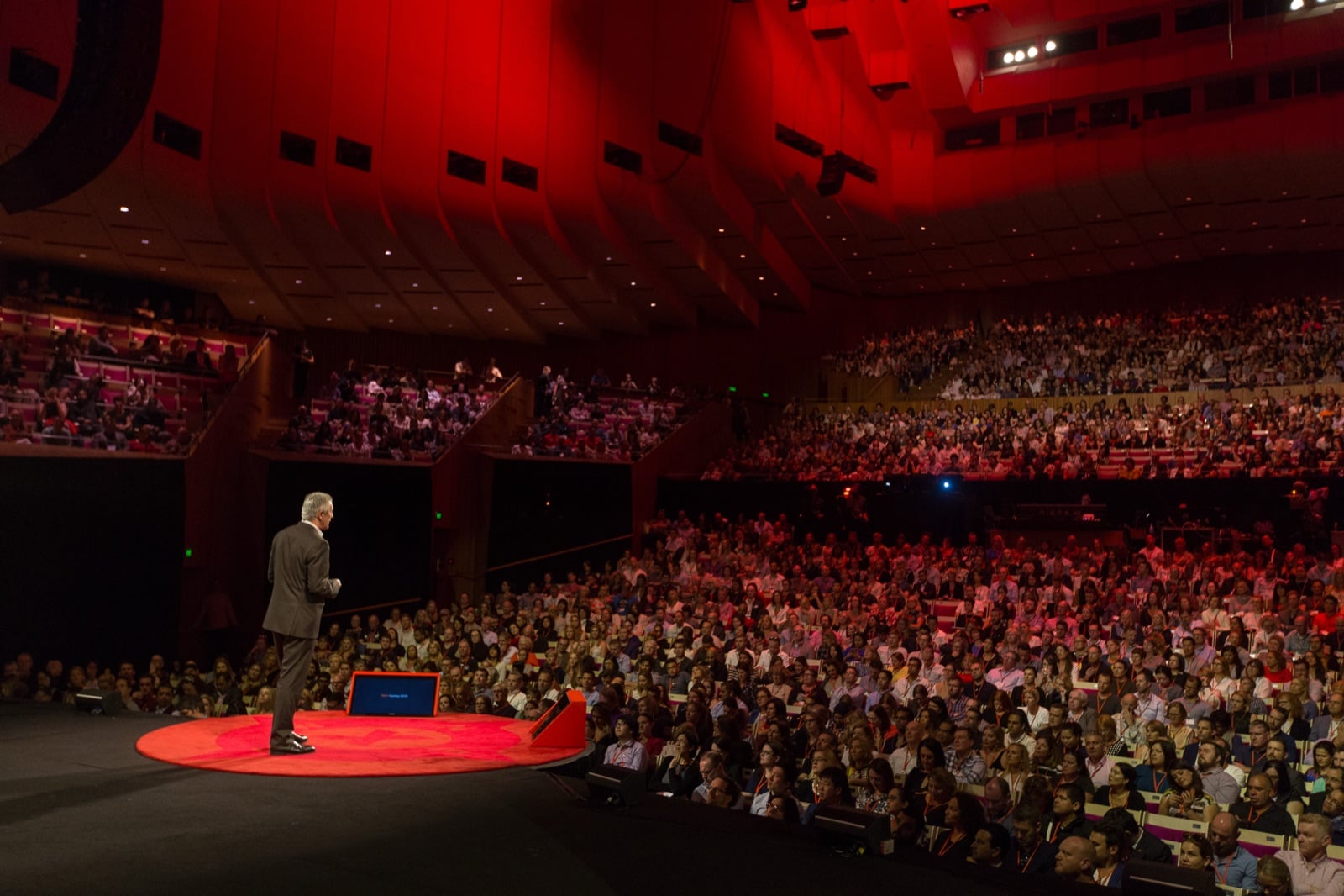 A speaker alone on the red disc at TEDxSydney, facing a packed concert hall